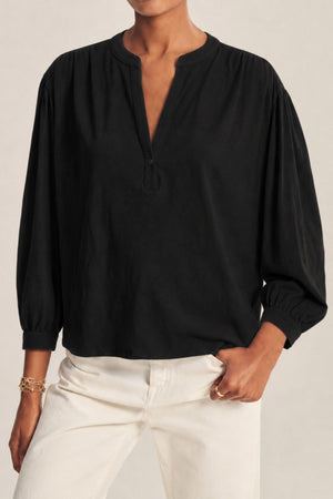 A person is wearing the KNOX TEE by Velvet by Graham & Spencer—a loose-fitting black long-sleeve blouse with a relaxed swing top silhouette and split neckline—paired with cream-colored pants and standing with one hand in a pocket.