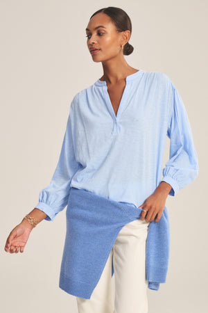 A woman in the KNOX TEE by Velvet by Graham & Spencer, a light blue cotton slub top, pairs it with white pants and a blue sweater tied at her waist, standing against a neutral background.