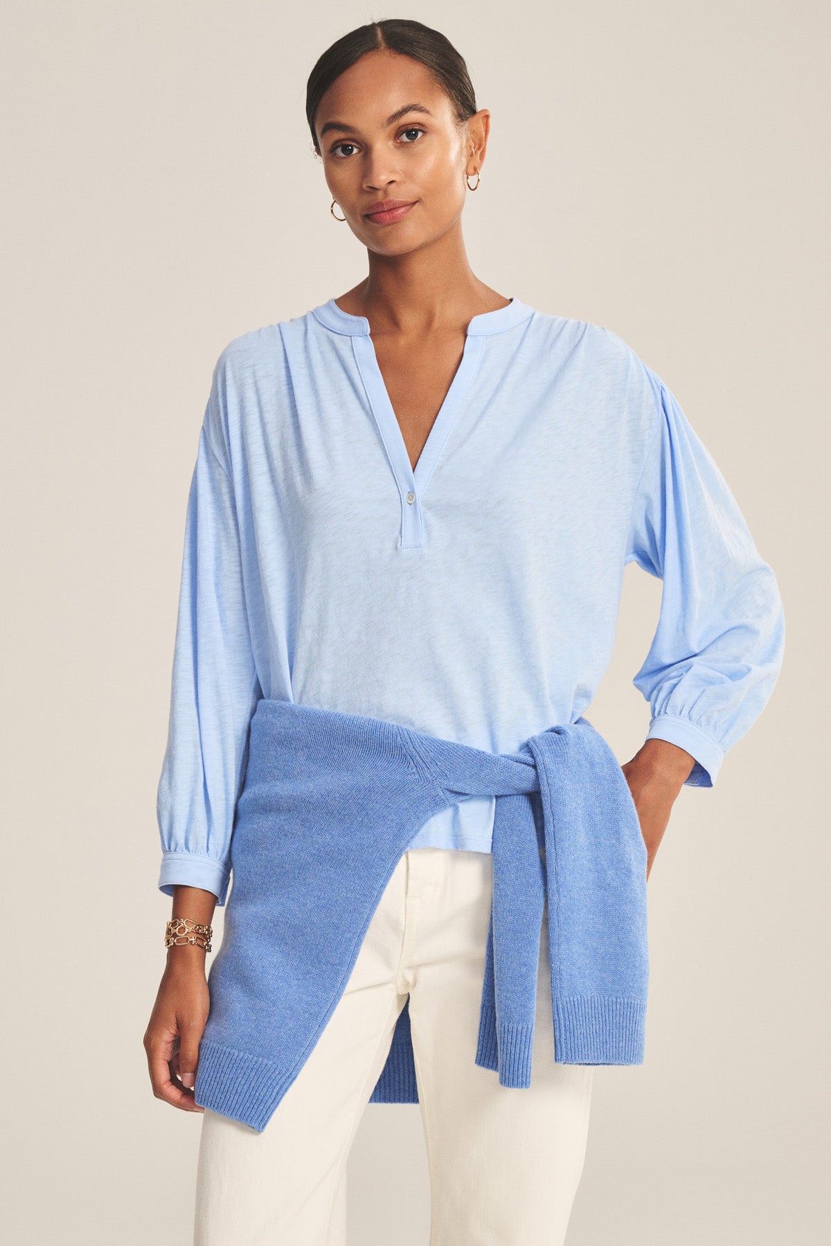 A woman faces the camera against a plain background wearing the KNOX TEE by Velvet by Graham & Spencer in light blue, white pants, and a blue sweater tied at her waist.