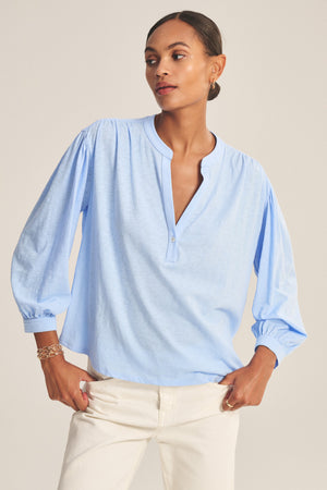 Model wearing the KNOX TEE by Velvet by Graham & Spencer—a light blue, relaxed V-neck long sleeve shirt—paired with white pants against a plain background.