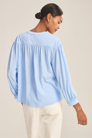 A woman with a low bun is seen from the back, wearing the Velvet by Graham & Spencer KNOX TEE in light blue and white pants against a neutral background.