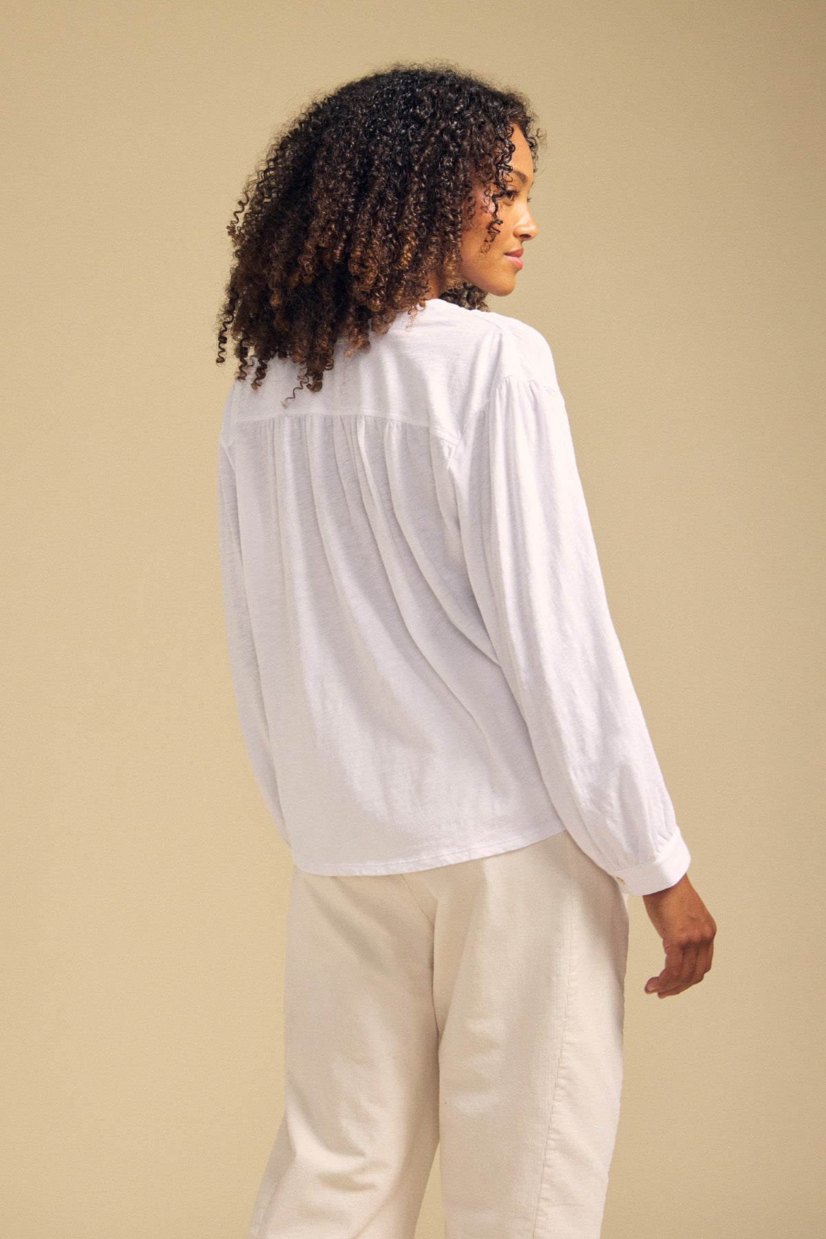 A person with curly hair wearing the KNOX TEE by Velvet by Graham & Spencer—a relaxed swing top with a V-neckline—paired with light pants stands facing away against a beige background.