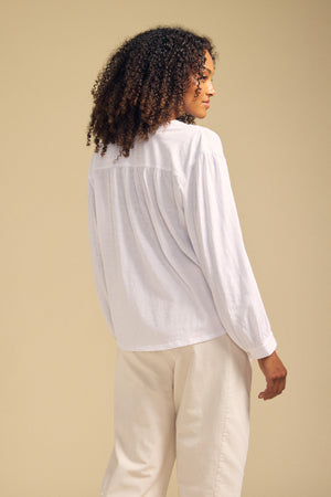 A person with curly hair wearing the KNOX TEE by Velvet by Graham & Spencer—a relaxed swing top with a V-neckline—paired with light pants stands facing away against a beige background.