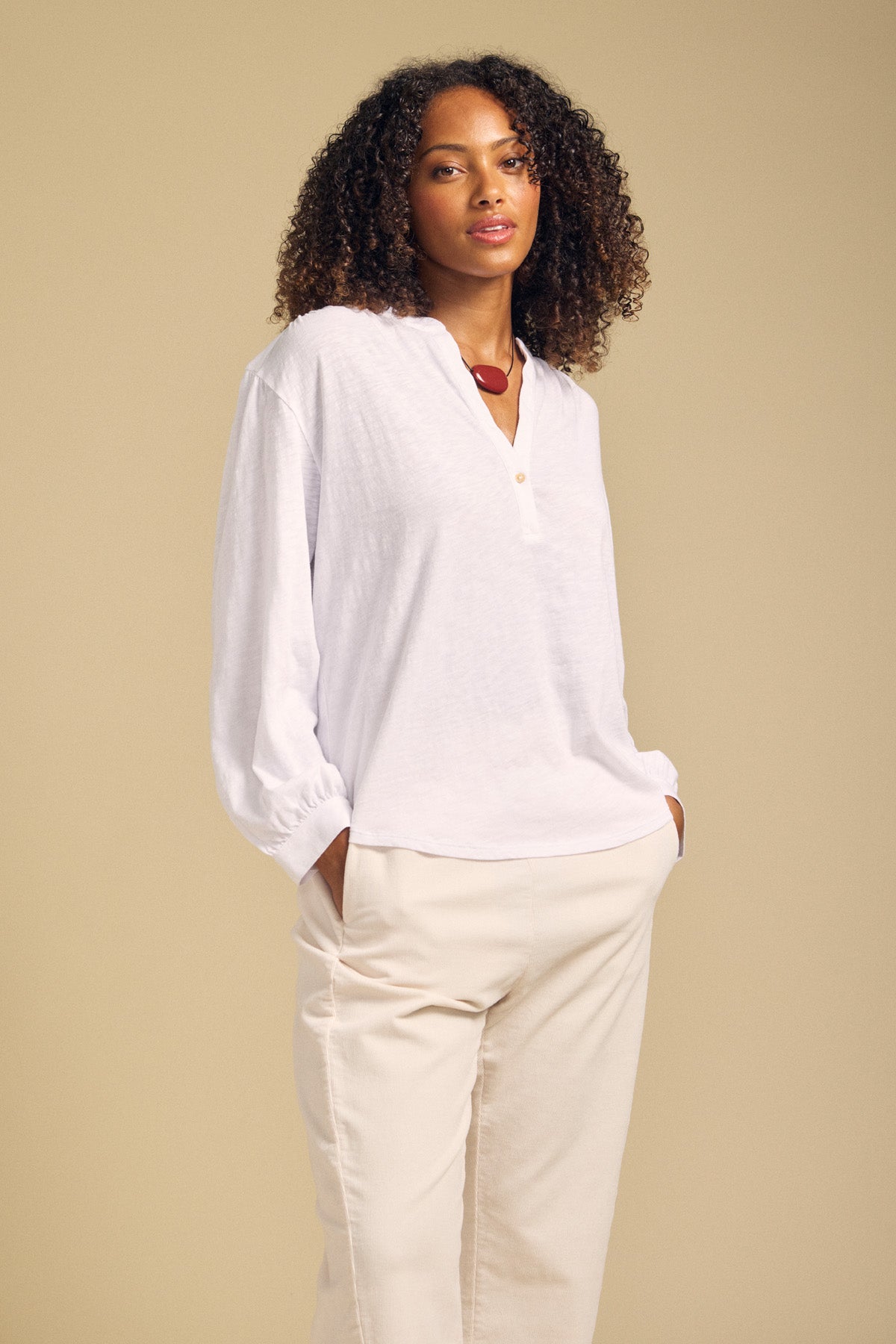 A woman stands with her hands in her pockets, wearing the relaxed KNOX TEE by Velvet by Graham & Spencer and light-colored pants against a beige background.-41929870082241