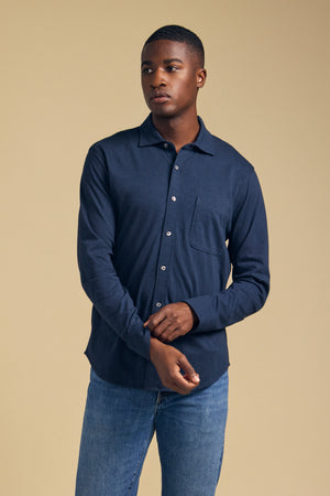 A man wearing the JONAH SHIRT by Velvet by Graham & Spencer—a navy blue long-sleeve button-up with a chest pocket—paired with blue jeans, stands against a beige background.