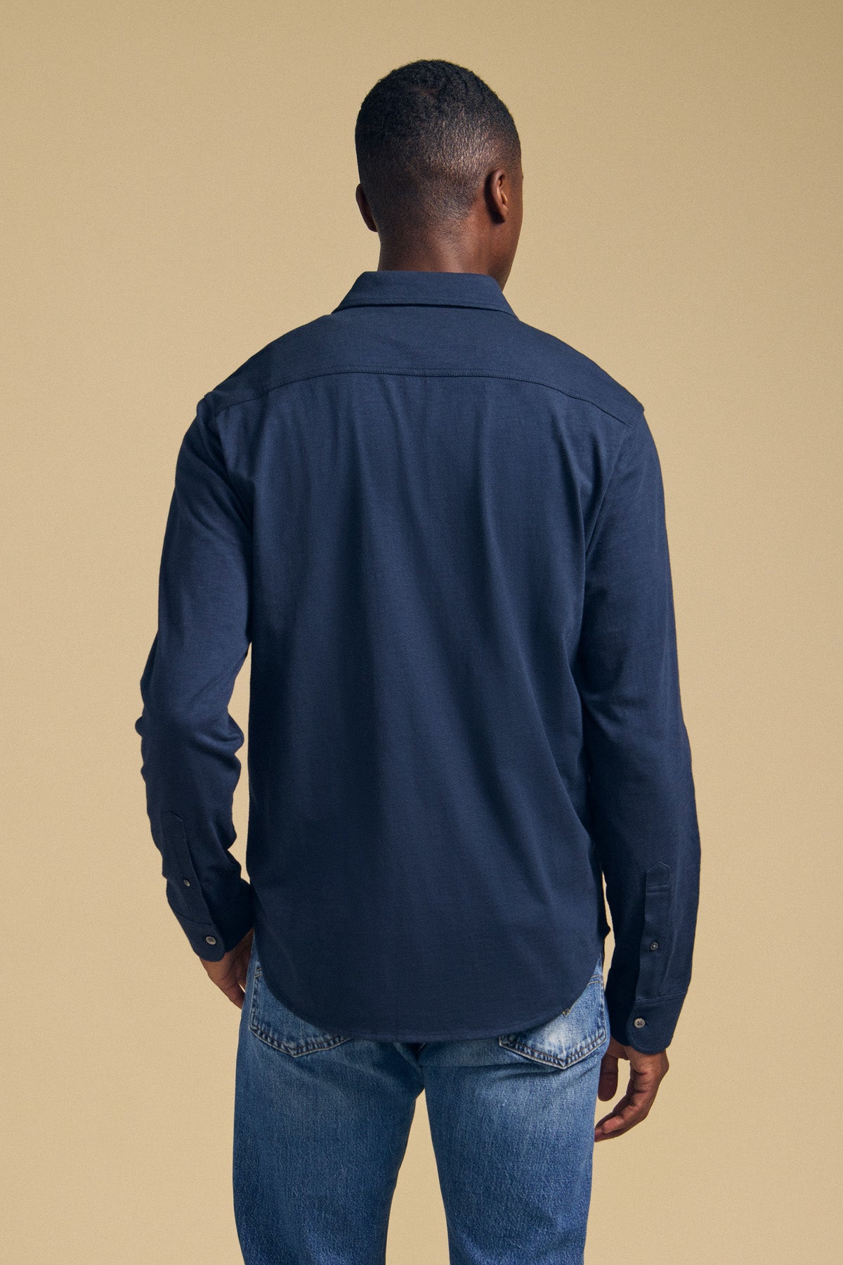 A person stands facing away from the camera, wearing the JONAH SHIRT by Velvet by Graham & Spencer—a dark blue long-sleeve shirt—paired with light blue jeans against a plain beige background.