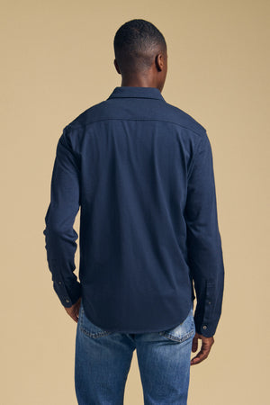 A person stands facing away from the camera, wearing the JONAH SHIRT by Velvet by Graham & Spencer—a dark blue long-sleeve shirt—paired with light blue jeans against a plain beige background.