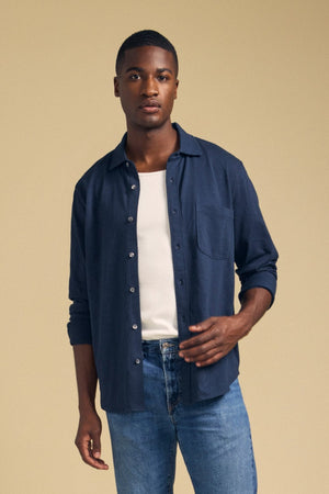 A person wearing the JONAH SHIRT by Velvet by Graham & Spencer, layered over a white t-shirt and paired with blue jeans, stands against a plain beige background.