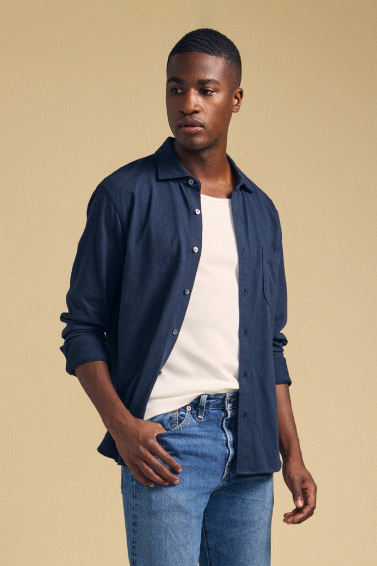 A man wearing the JONAH SHIRT by Velvet by Graham & Spencer over a white t-shirt and blue jeans stands against a beige background.