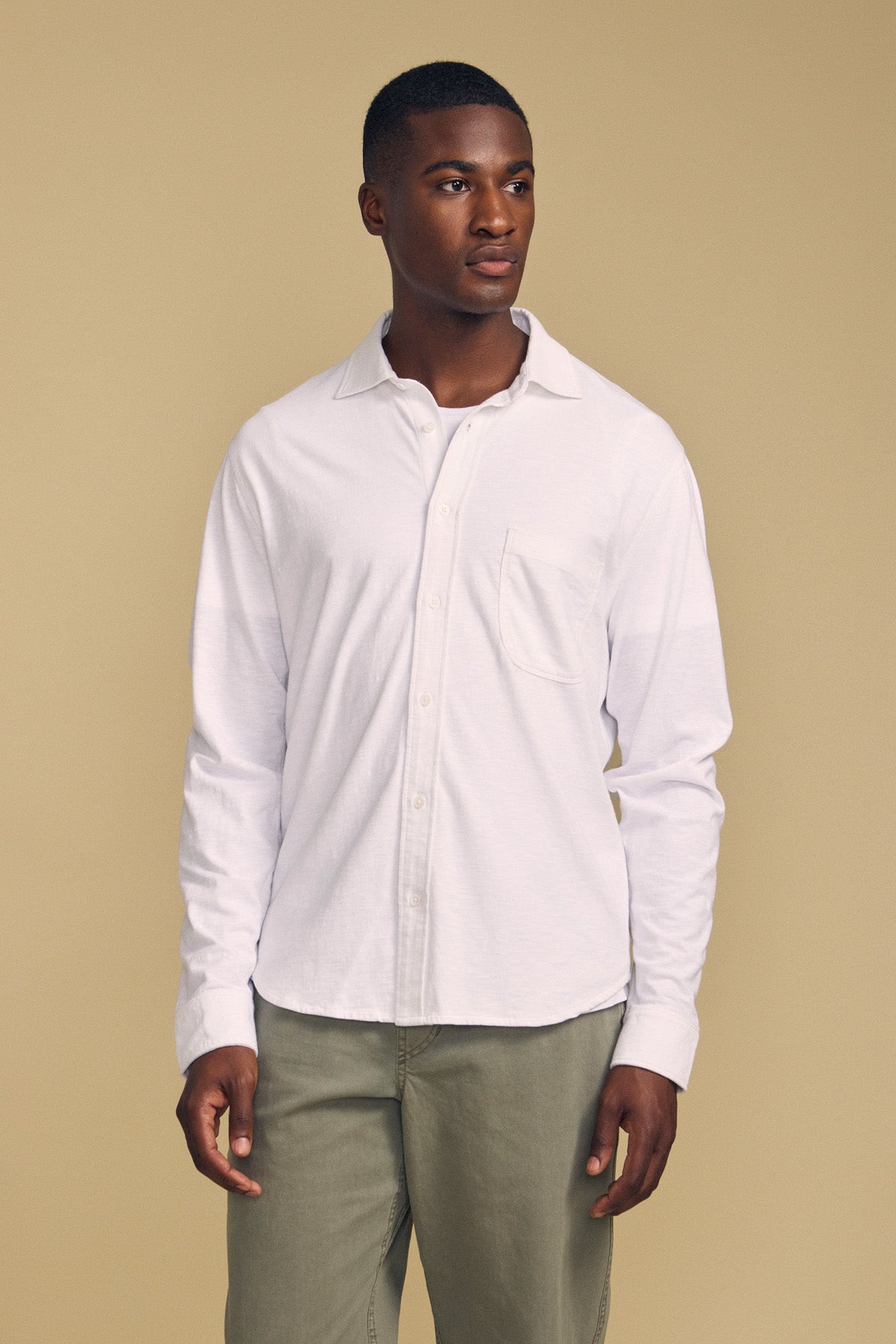 A man stands against a beige background, wearing the JONAH SHIRT by Velvet by Graham & Spencer—a white long-sleeve button-up—paired with light olive green pants.-41859679256769