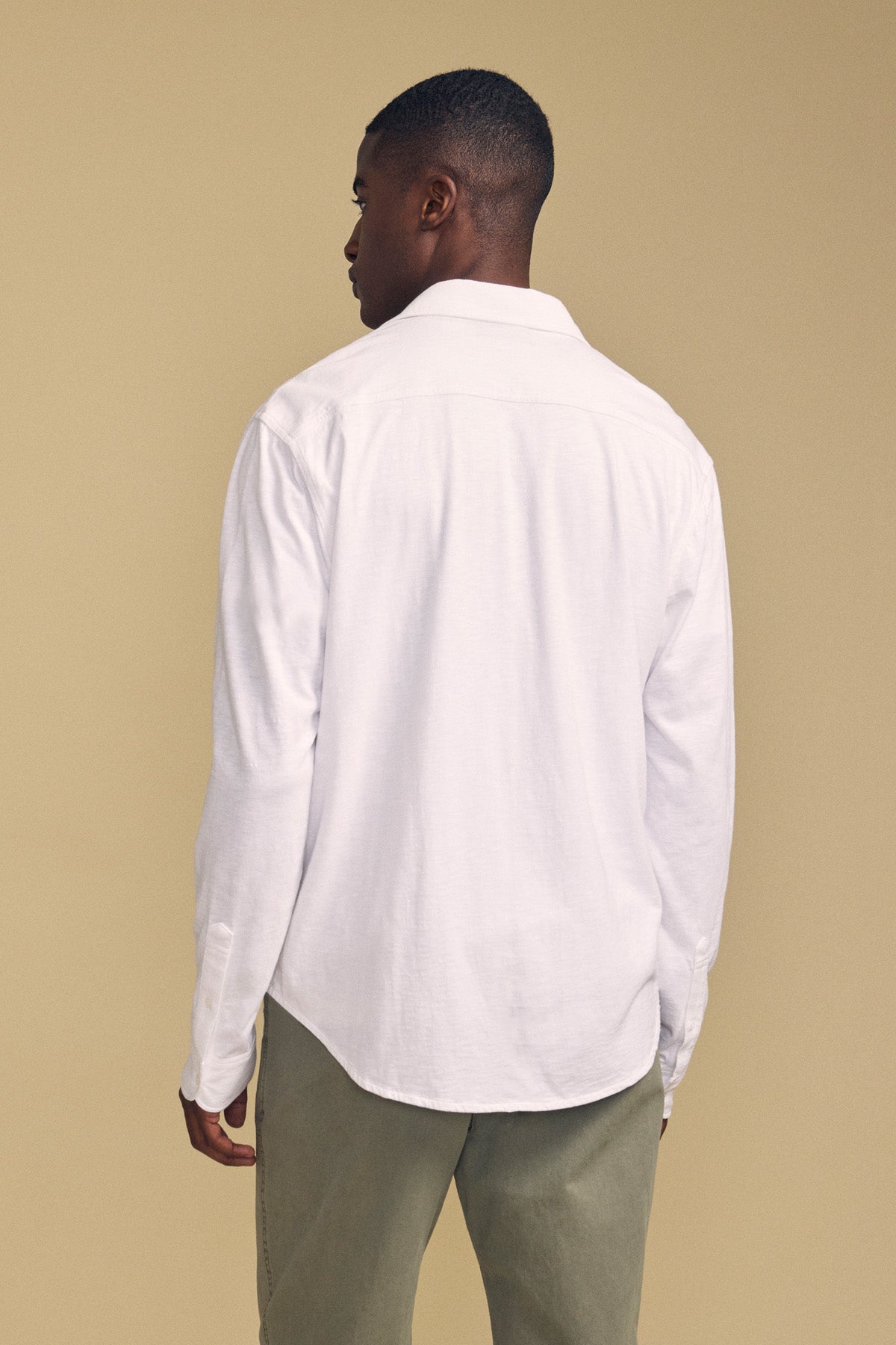 A person stands facing away from the camera against a plain beige background, wearing the JONAH SHIRT by Velvet by Graham & Spencer—a long-sleeve white button-up shirt—paired with olive green pants.-41859679748289