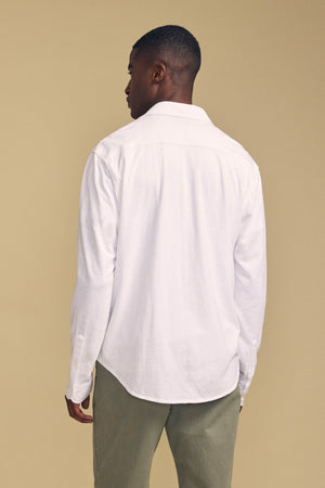 A person stands facing away from the camera against a plain beige background, wearing the JONAH SHIRT by Velvet by Graham & Spencer—a long-sleeve white button-up shirt—paired with olive green pants.
