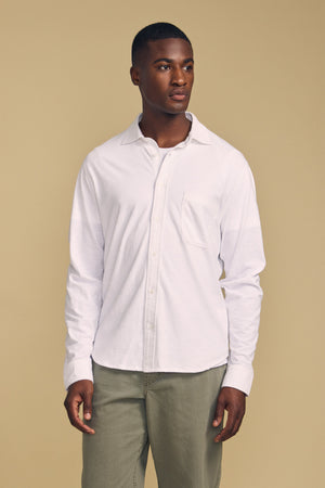 A man stands against a beige background, wearing the JONAH SHIRT by Velvet by Graham & Spencer—a white long-sleeve button-up—paired with light olive green pants.