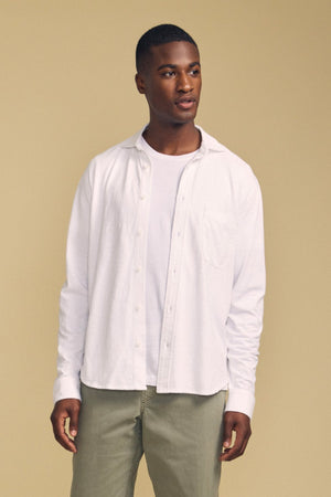 A man stands against a beige background wearing the JONAH SHIRT by Velvet by Graham & Spencer—a white button-up shirt—layered over a white t-shirt and paired with light green pants.