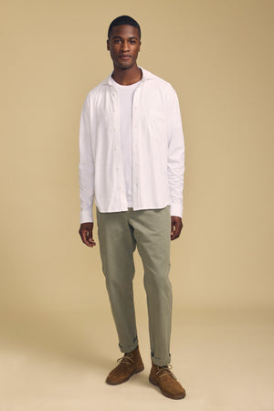A man stands against a beige background, wearing the JONAH SHIRT by Velvet by Graham & Spencer over a white t-shirt, paired with light green pants and brown suede shoes.