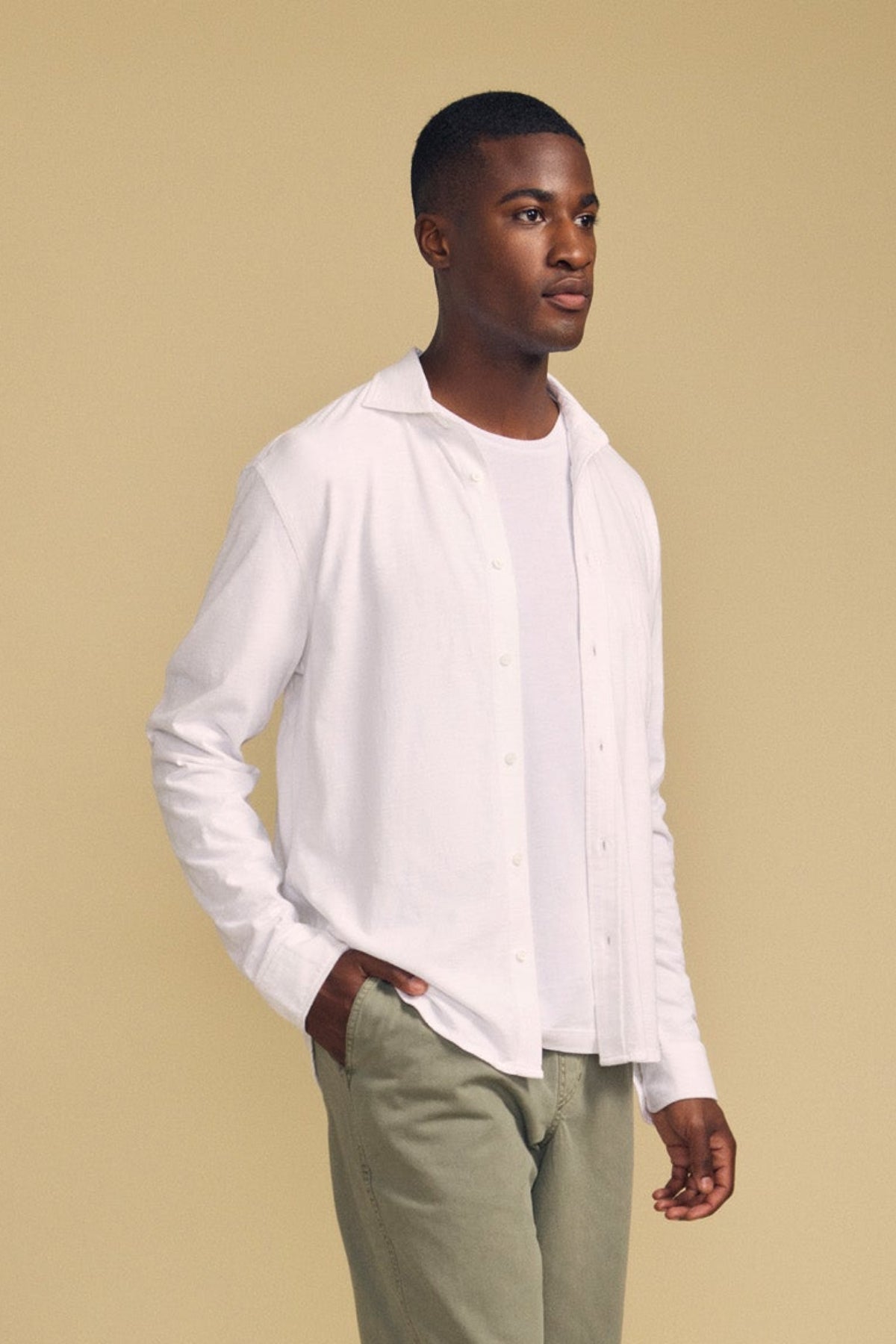 A person stands against a beige background, wearing the JONAH SHIRT by Velvet by Graham & Spencer—layered over a white t-shirt and paired with light olive-green pants, with one hand in their pocket.-41859679486145