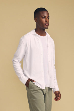 A person stands against a beige background, wearing the JONAH SHIRT by Velvet by Graham & Spencer—layered over a white t-shirt and paired with light olive-green pants, with one hand in their pocket.