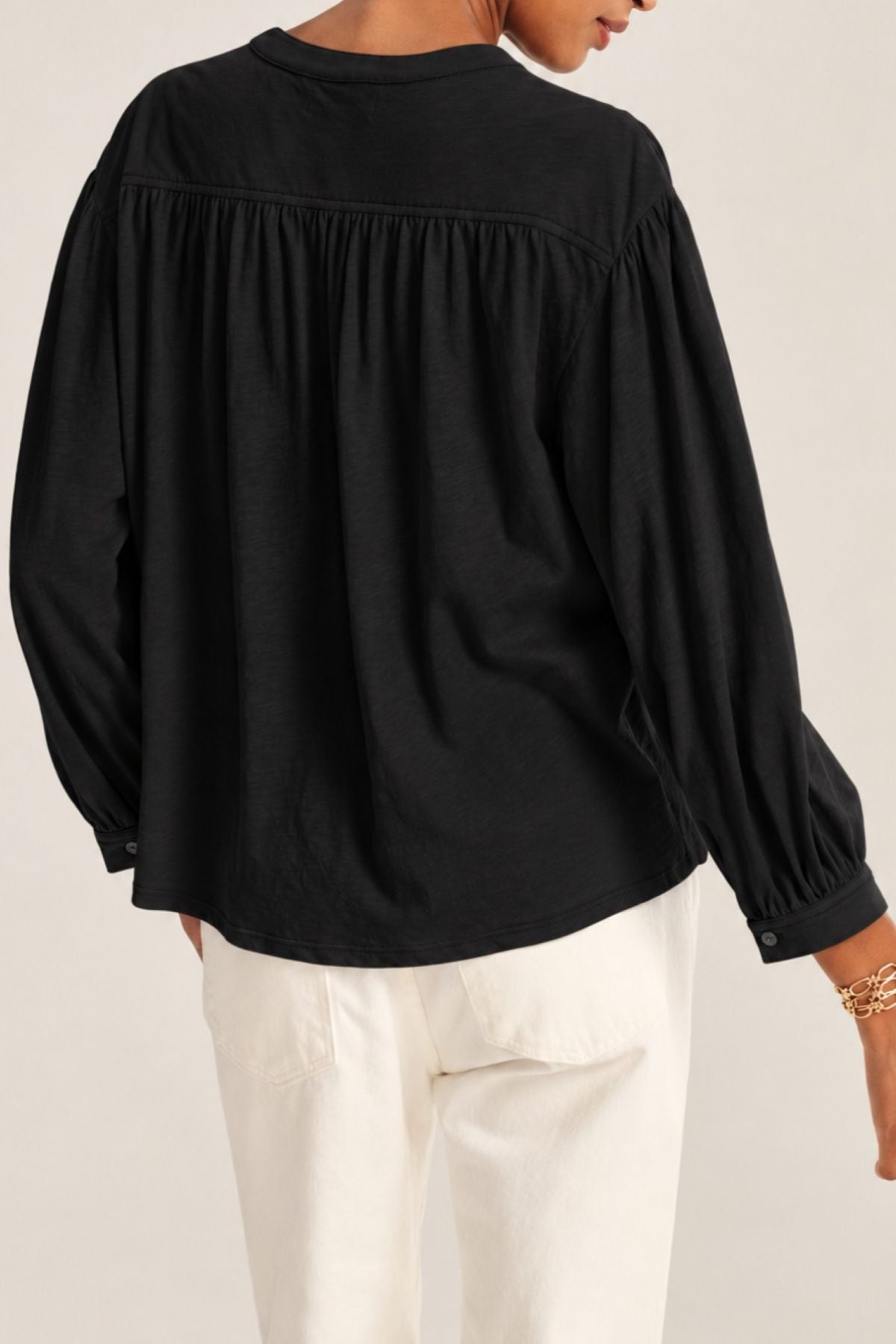 A person wearing the Velvet by Graham & Spencer KNOX TEE—a loose-fitting black swing-style top with gathered back detail—and white pants, shown from behind.-42059273961665