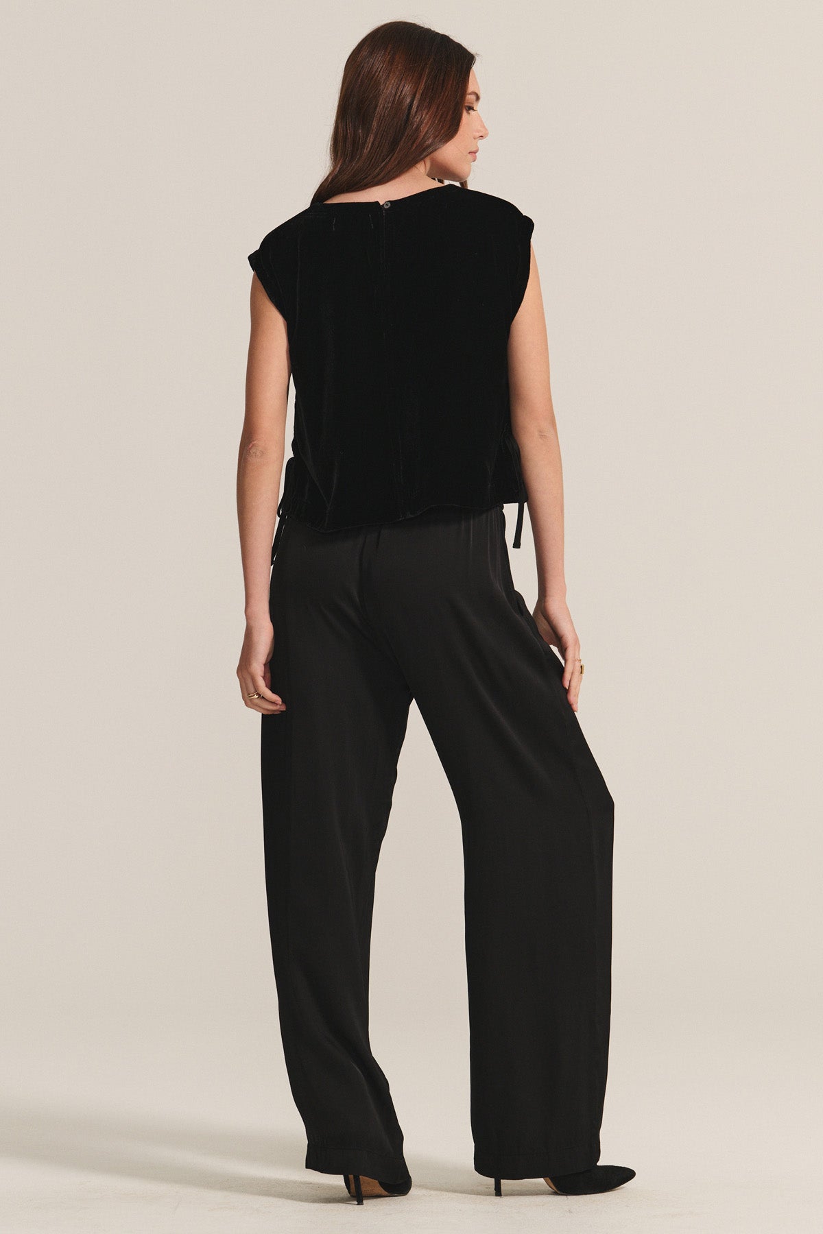   A woman stands facing away in the LARKIN PANT by Velvet by Graham & Spencer, paired with a sleeveless black velvet top and black heels—a flattering, everyday look shown against a plain light background. 