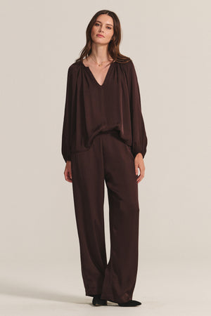 A woman stands against a plain background, wearing a loose-fitting, long-sleeved dark brown blouse and the LARKIN PANT by Velvet by Graham & Spencer—wide-leg satin pants paired with black shoes for effortless everyday style.