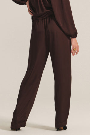 A person wears the LARKIN PANT by Velvet by Graham & Spencer—loose-fitting, dark brown wide-leg satin pants styled with a matching long-sleeve top for a flattering fit and stylish everyday wear.