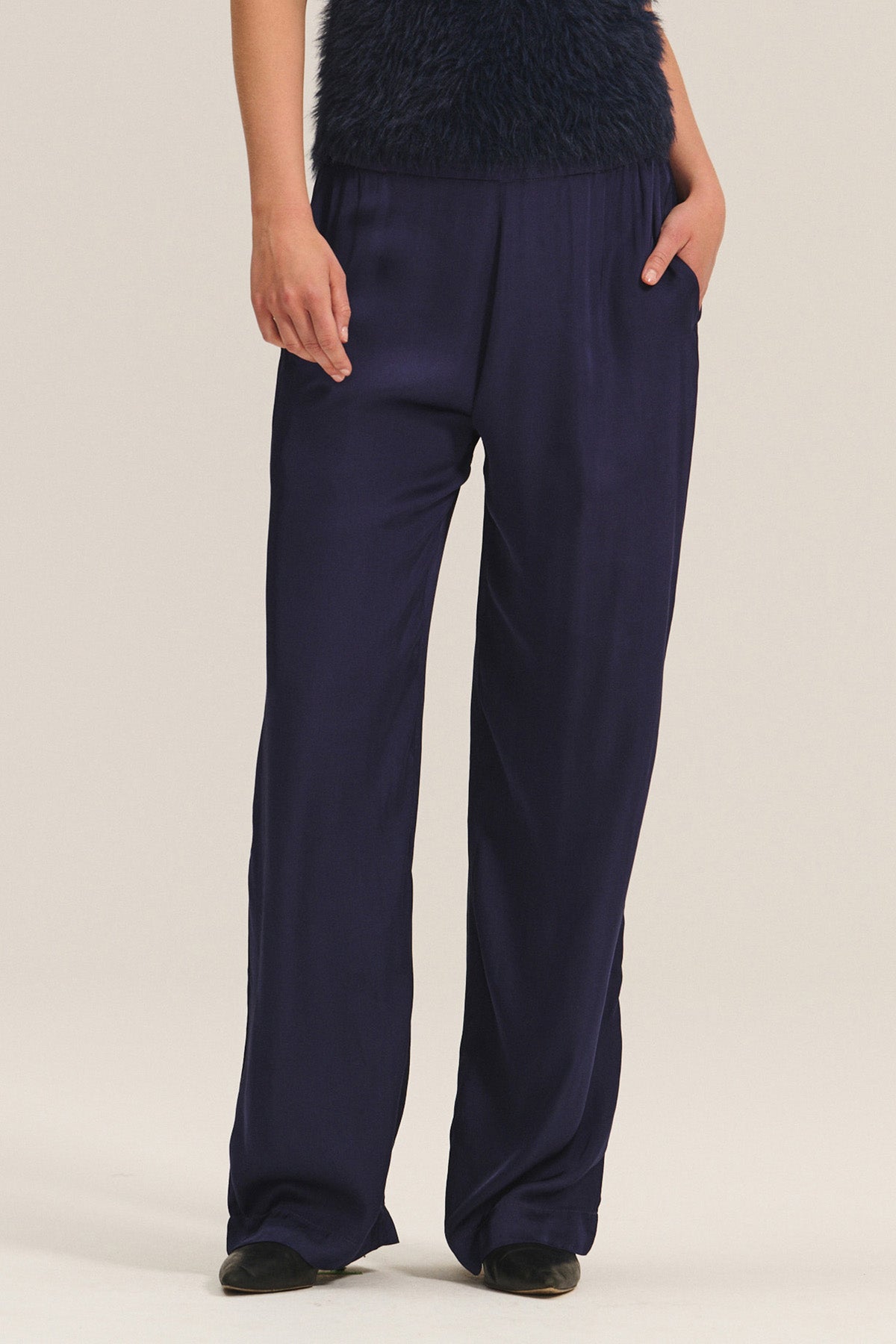 Model showcases the Velvet by Graham & Spencer LARKIN PANT in dark navy satin, styled with a textured top and black shoes. The wide-leg, flattering fit offers everyday comfort. Only the lower half of the body is visible; hands in pockets.-40263653032129