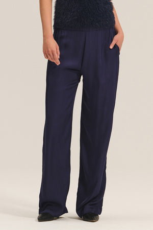 Model showcases the Velvet by Graham & Spencer LARKIN PANT in dark navy satin, styled with a textured top and black shoes. The wide-leg, flattering fit offers everyday comfort. Only the lower half of the body is visible; hands in pockets.