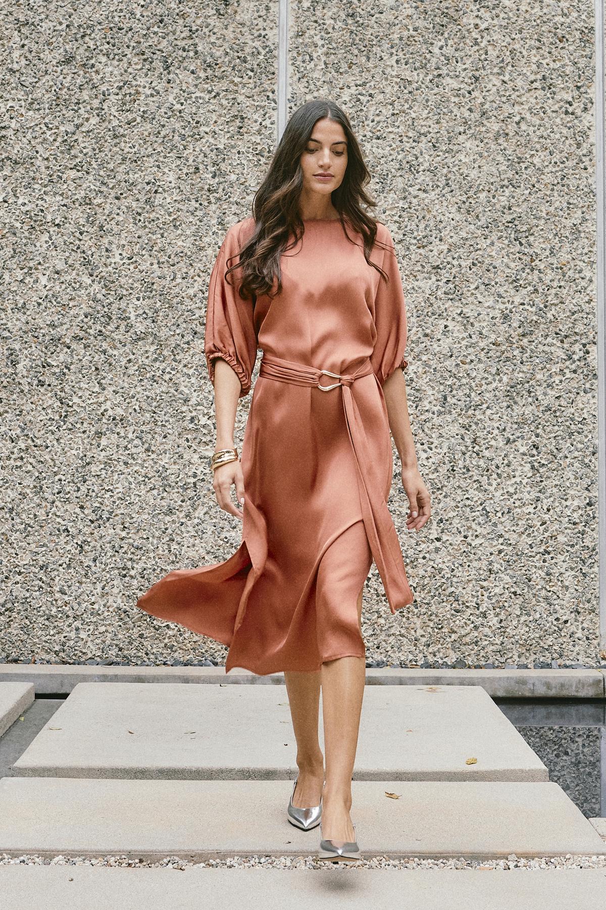   A woman in the LAUREL SATIN DRESS by Velvet by Graham & Spencer, showcasing elegant dolman sleeves, walks on a concrete path against a textured wall. 