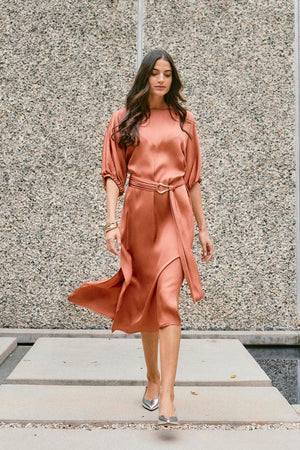 A woman in the LAUREL SATIN DRESS by Velvet by Graham & Spencer, showcasing elegant dolman sleeves, walks on a concrete path against a textured wall.