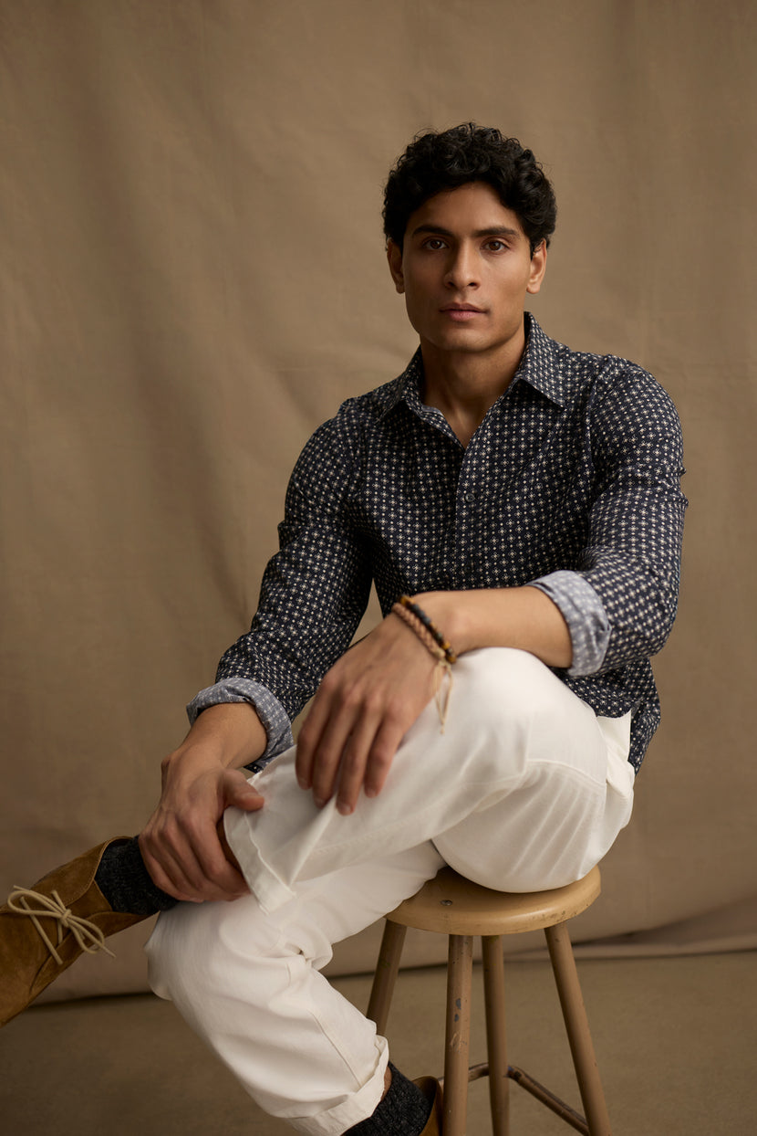 A man with short dark hair sits on a wooden stool against a plain brown backdrop, wearing the LAWRENCE BUTTON-UP SHIRT by Velvet by Graham & Spencer in breathable cotton, styled with white pants and brown shoes for a tailored look.
