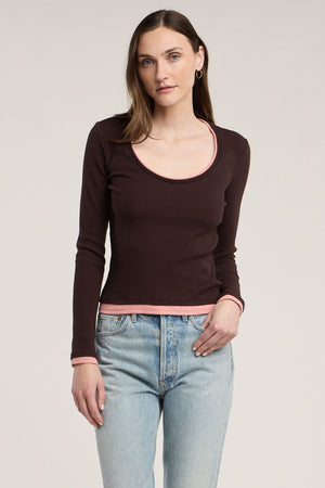 A woman wears the MALANI TEE by Velvet by Graham & Spencer—a long-sleeve, dark brown ribbed top with pink trim—paired with light blue jeans and set against a plain light background. Perfect for layering, this stretch cotton piece effortlessly combines comfort and style.