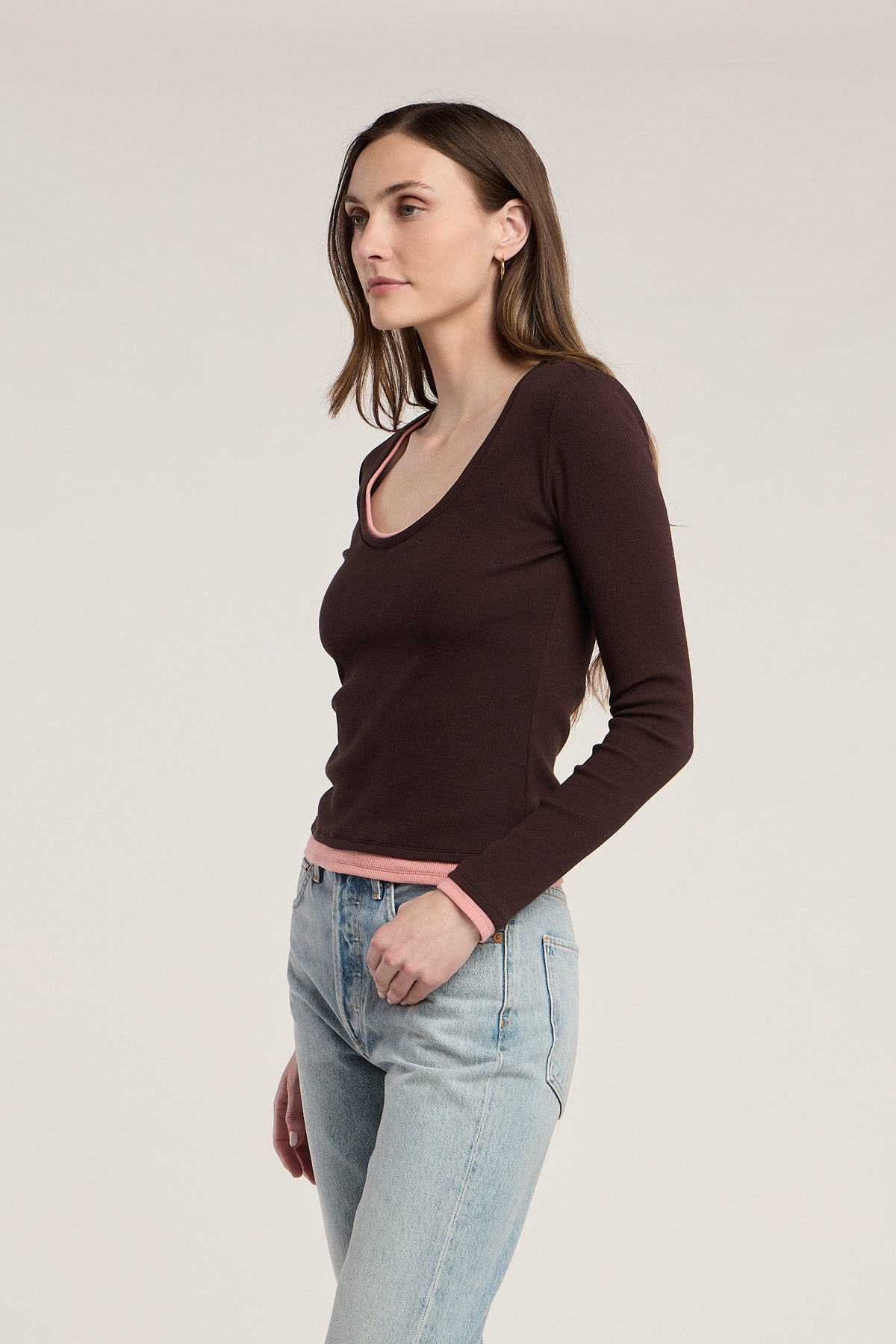 A woman stands facing left, wearing the MALANI TEE by Velvet by Graham & Spencer—a dark ribbed top with pink trim—paired with light blue jeans against a plain background, perfect for effortless layering.-42063810035905