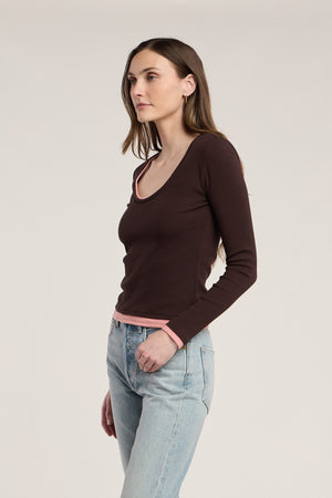 A woman stands facing left, wearing the MALANI TEE by Velvet by Graham & Spencer—a dark ribbed top with pink trim—paired with light blue jeans against a plain background, perfect for effortless layering.