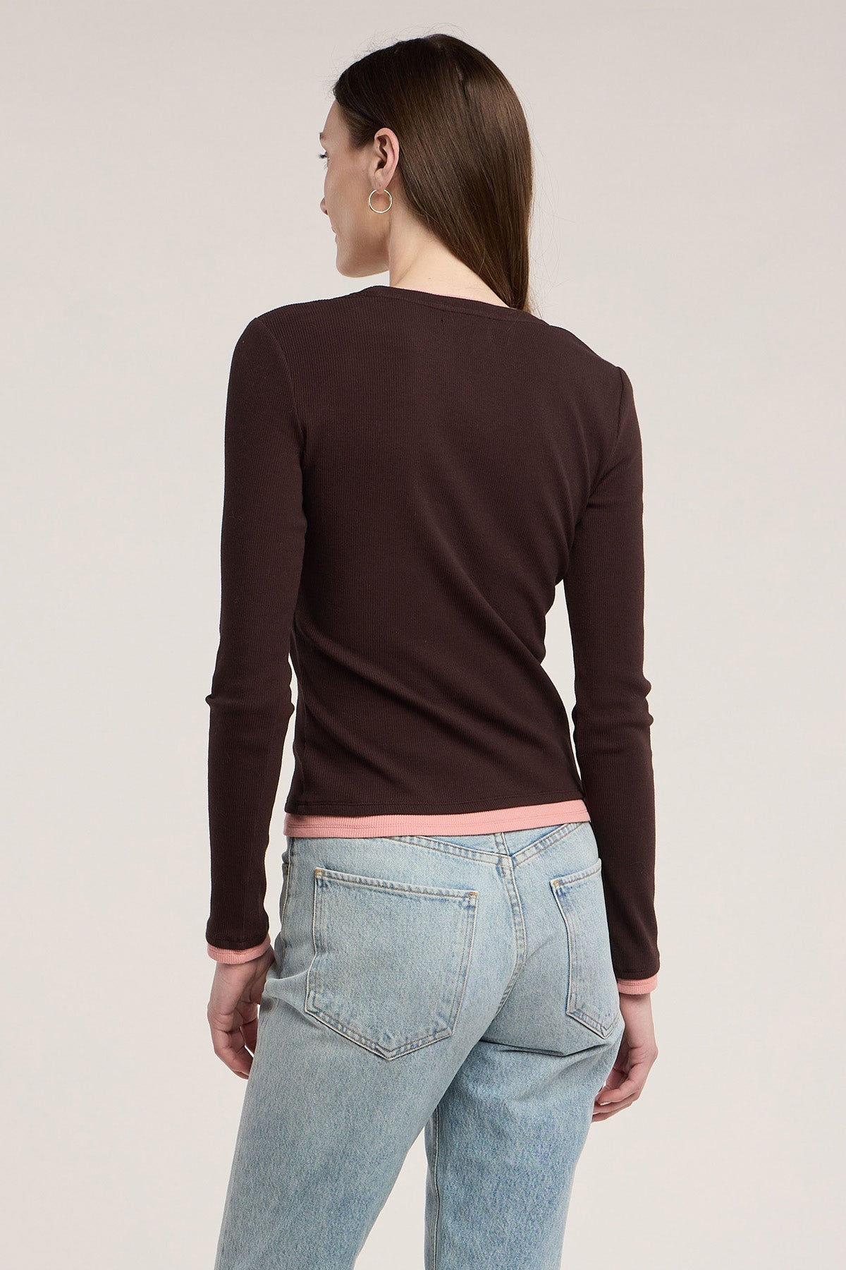 Wearing the Velvet by Graham & Spencer MALANI TEE—a long-sleeve dark brown ribbed top with pink trim—paired with light blue jeans, this person stands facing away against a plain background, showcasing an ideal layering look.