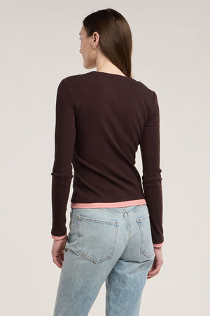 Wearing the Velvet by Graham & Spencer MALANI TEE—a long-sleeve dark brown ribbed top with pink trim—paired with light blue jeans, this person stands facing away against a plain background, showcasing an ideal layering look.