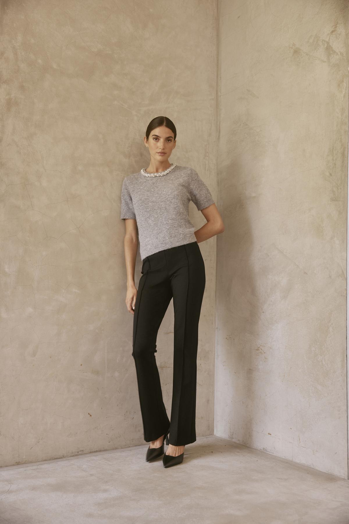   A woman wearing the LAYTON ALPACA EMBELLISHED SWEATER by Velvet by Graham & Spencer and black pants stands against a textured beige wall. 