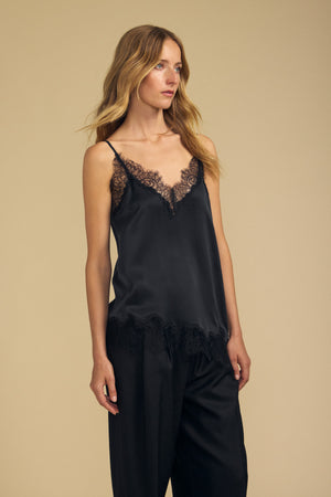 A woman wearing the BEVERLY SILK LACE CAMISOLE by Velvet by Graham & Spencer—a black silk camisole with lace trim and adjustable straps—paired with matching loose black pants, stands against a plain beige background.