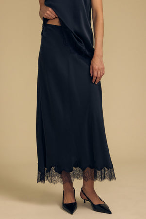 Wearing the KRISTIN SILK SKIRT by Velvet by Graham & Spencer—featuring black silk, an elegant lace trim, and a comfortable elastic waist—a person stands against a beige background, paired with black pointed-toe heels.