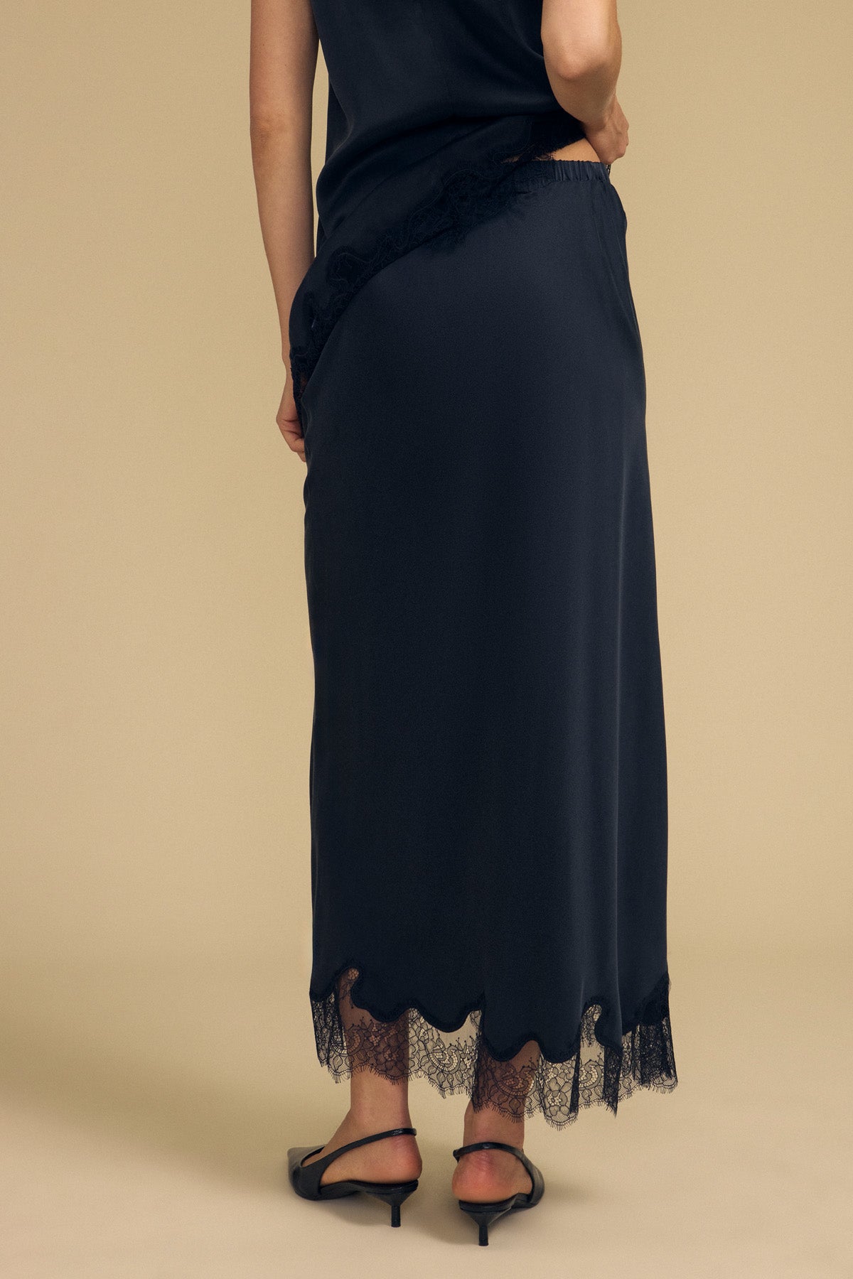 A person wearing the KRISTIN SILK SKIRT by Velvet by Graham & Spencer—featuring black silk fabric with lace trim—and black heels, stands on a beige background, photographed from the back.