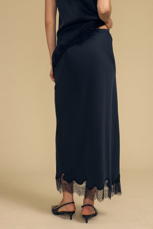 A person wearing the KRISTIN SILK SKIRT by Velvet by Graham & Spencer—featuring black silk fabric with lace trim—and black heels, stands on a beige background, photographed from the back.