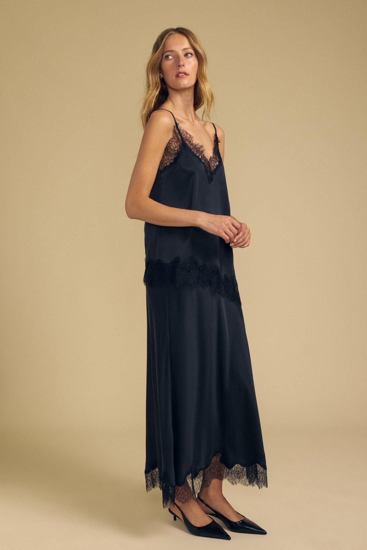 A woman stands in a studio against a beige background, wearing the KRISTIN SILK SKIRT by Velvet by Graham & Spencer. The black silk skirt features an elegant elastic waist that gently cinches her silhouette and is styled with delicate lace trim and black pointed heels.
