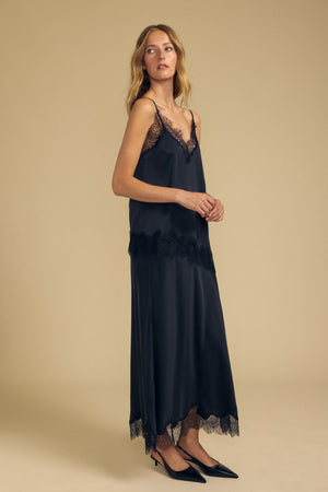 A woman stands in a studio against a beige background, wearing the KRISTIN SILK SKIRT by Velvet by Graham & Spencer. The black silk skirt features an elegant elastic waist that gently cinches her silhouette and is styled with delicate lace trim and black pointed heels.