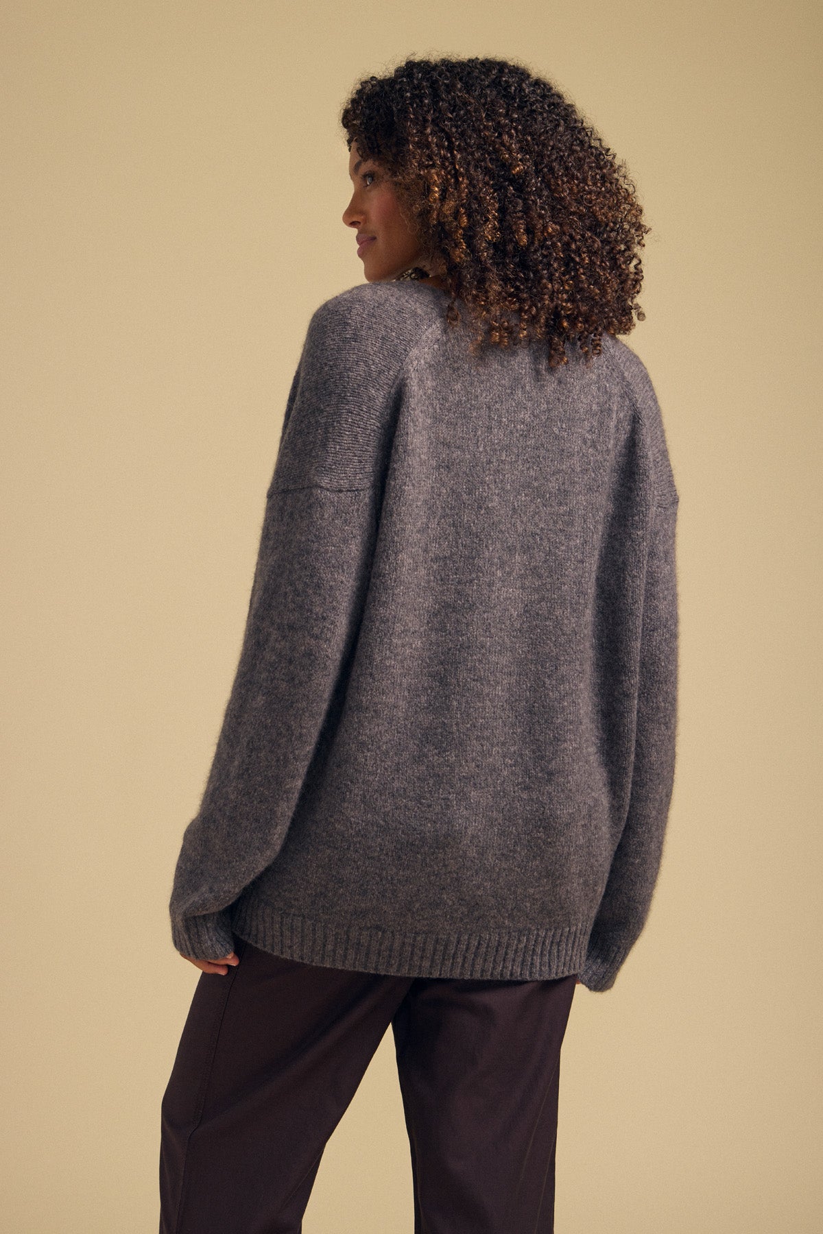 Wearing the LEA CARDIGAN by Velvet by Graham & Spencer—a cozy, loose gray knit—this person with curly hair pairs it with dark pants and stands facing away from the camera against a plain beige background, creating an effortless look that’s perfect for everyday layering.