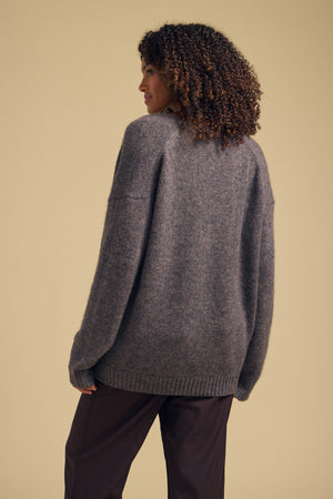 Wearing the LEA CARDIGAN by Velvet by Graham & Spencer—a cozy, loose gray knit—this person with curly hair pairs it with dark pants and stands facing away from the camera against a plain beige background, creating an effortless look that’s perfect for everyday layering.