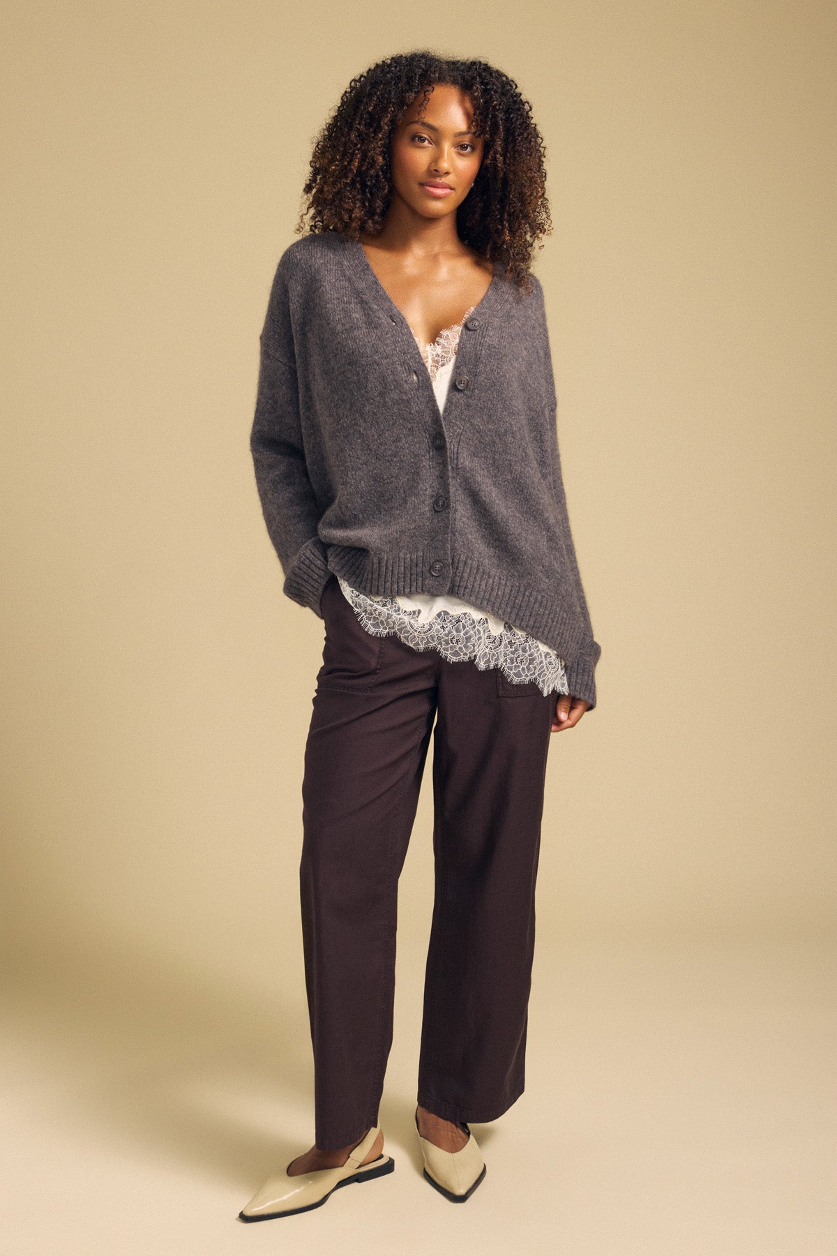 A woman stands in a studio with her hands in her pockets, wearing the LEA CARDIGAN by Velvet by Graham & Spencer—a cozy wool-cashmere layer—paired with a white lace top, dark brown pants, and cream-colored shoes against a beige background for an effortlessly stylish everyday look.