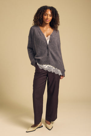 A woman stands in a studio with her hands in her pockets, wearing the LEA CARDIGAN by Velvet by Graham & Spencer—a cozy wool-cashmere layer—paired with a white lace top, dark brown pants, and cream-colored shoes against a beige background for an effortlessly stylish everyday look.