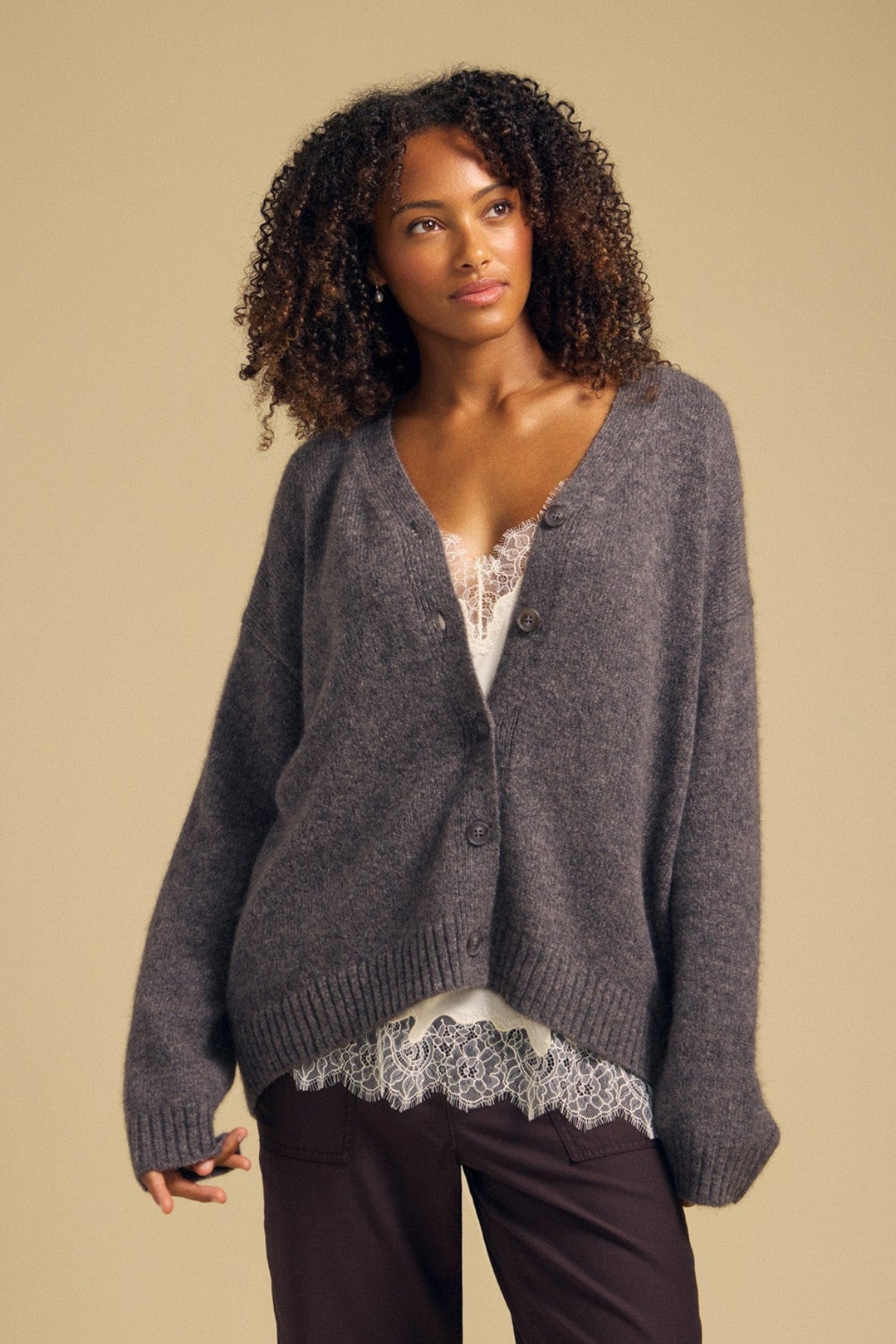 A woman stands against a plain beige background, effortlessly layering the Velvet by Graham & Spencer LEA CARDIGAN—a cozy wool-cashmere piece—over a white lace-trimmed top and dark pants.