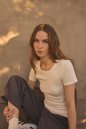 A woman with long brown hair sits on a chair against a beige wall, wearing the LENA POINTELLE TEE by Velvet by Graham & Spencer—a white cotton knit top with a fitted silhouette—paired with gray pants and white socks with sneakers. Warm light creates soft shadows on the wall.