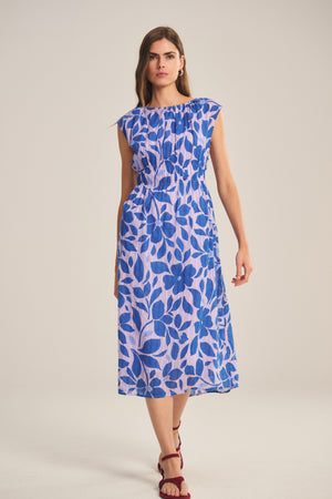 A woman wears the LEVAINE DRESS by Velvet by Graham & Spencer, a sleeveless blue and white floral print cotton dress with a smocked waist, paired with red strappy sandals, standing against a plain background.