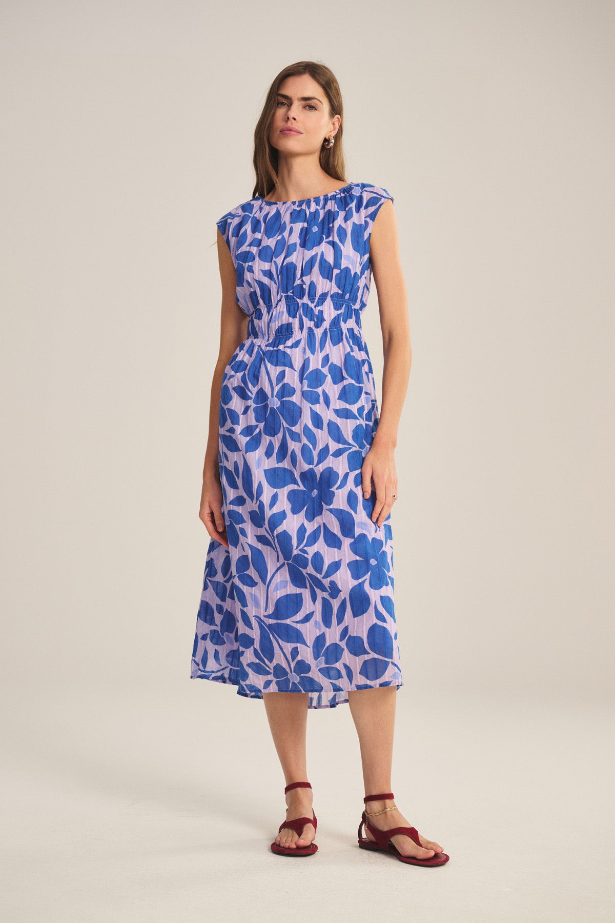 A woman wears the LEVAINE DRESS by Velvet by Graham & Spencer—a blue and white floral smocked waist dress with cap sleeves—paired with red flat sandals, standing against a plain light background.